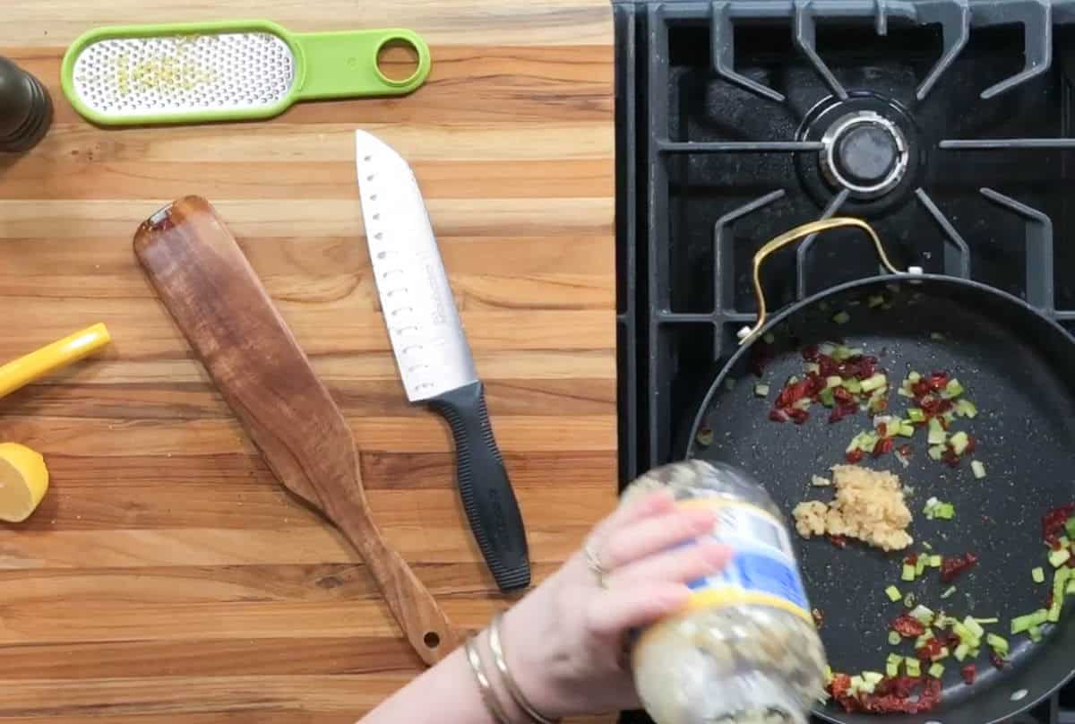 A hand holding an open jar of minced garlic over a frying pan with chopped ingredients. A knife, wooden spatula, and grater are on the wooden countertop near a stovetop with a black pan.