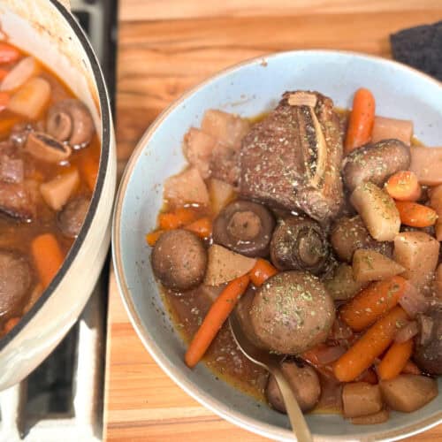 A ceramic bowl filled with a beef stew containing chunks of beef, whole mushrooms, baby carrots, and potatoes, garnished with green herbs. Beside it, a pot on a wooden surface holds more stew of the same ingredients. A spoon rests in the bowl.