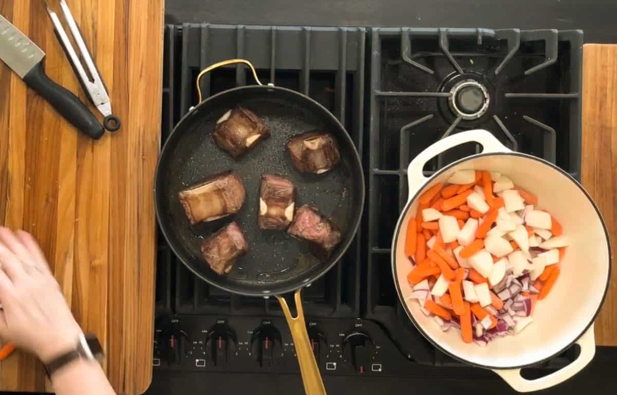A stove with two pans. The left pan has four pieces of browned meat, possibly short ribs. The right is a white pot containing chopped carrots, onions, and potatoes. Part of a person's hand is visible to the left, near a knife on a wooden countertop.