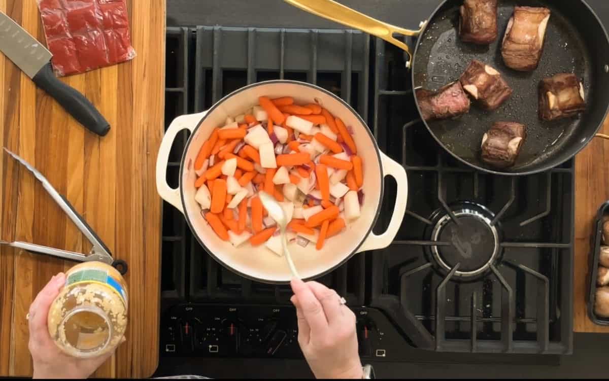 A person is stirring chopped carrots and onions in a white pot on a stove. Beside the pot, short ribs are being seared in a pan. Garlic in a jar, a knife, and a pair of tongs are on the wooden countertop.
