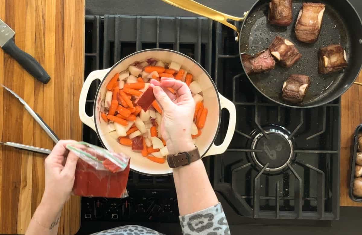 A person wearing a leopard print top adds tomato paste to a pot filled with chopped carrots and onions on a stovetop. A skillet with browned meat pieces is on the side, and a bag of tomato paste is held in one hand.