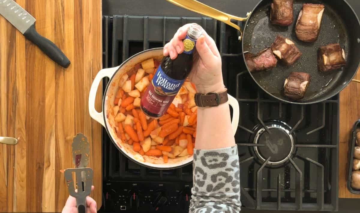 A person holding a bottle of red wine above a pot filled with sliced carrots and potatoes on a stovetop. A frying pan with seared meat pieces is on the adjacent burner. A spatula and knife rest on a wooden cutting board nearby.