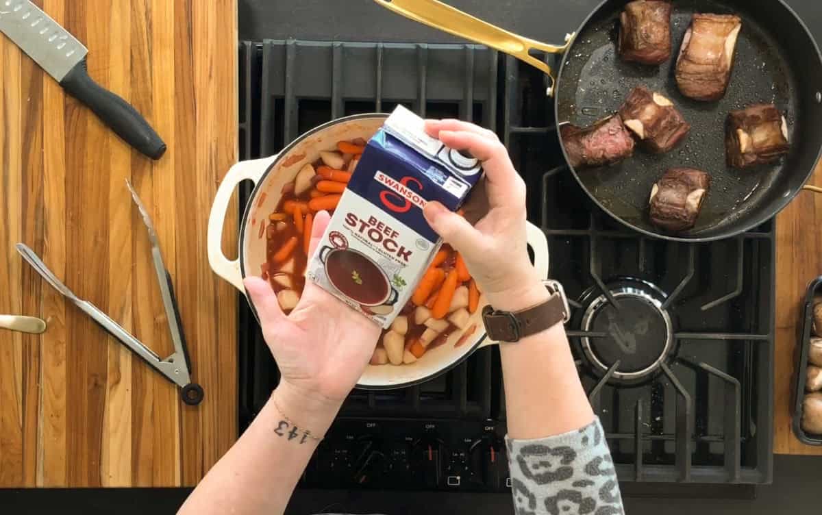A person holds a carton of beef stock over a pot filled with carrots, onions, and potatoes on a stovetop. A separate pan contains several browned pieces of meat. Kitchen utensils, including a knife and tongs, are on the wooden counter.