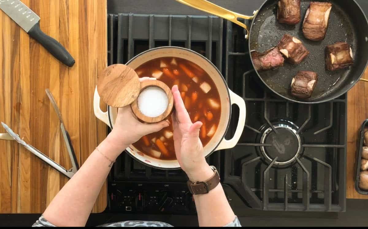 A person is adding salt from a wooden container to a pot of stew containing vegetables on a stove. Next to the pot is a pan with seared meat pieces. The scene includes kitchen utensils on a wooden cutting board.