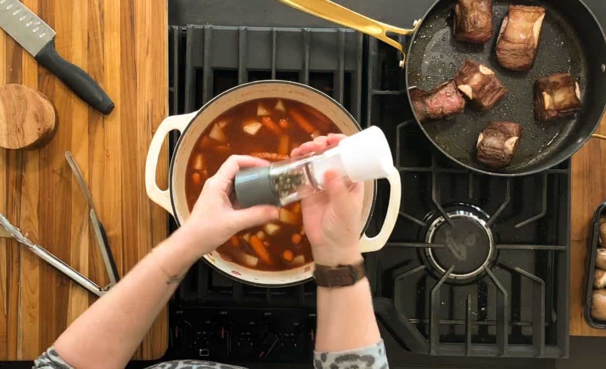 Overhead view of a person seasoning a pot of stew on a stovetop. Next to the pot is a frying pan with browned meat. A wooden cutting board with a knife and other utensils is on the side. The stove has a modern, black design.