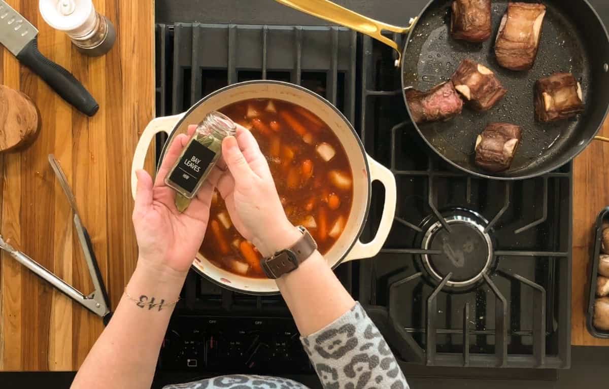 A person is adding a jar of bay leaves to a pot containing a stew with carrots and onions. Next to it, a pan with several cooked beef pieces is on the stove. A wooden cutting board with kitchen tools is nearby.