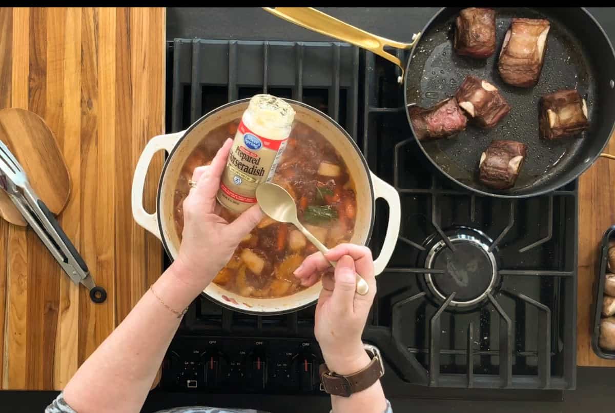 A person is adding a teaspoon of horseradish from a jar into a pot of stew on a stovetop. Next to it, a pan holds several pieces of browned meat. The countertop is wooden, and a pair of tongs is placed nearby.