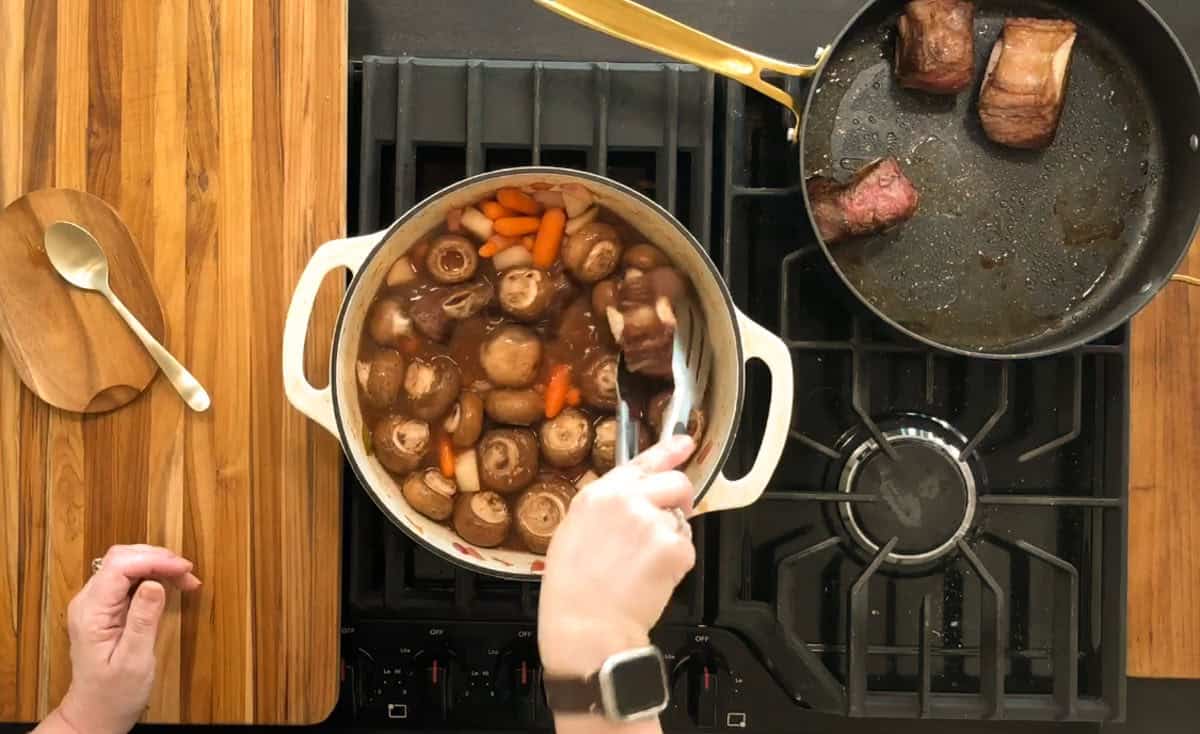 A person holding a pot on a stove, stirring a mixture of mushrooms, carrots, and sauce with tongs. A skillet with meat pieces is on the right. A wooden spoon rests on a small board to the side. A cutting board is on the left.