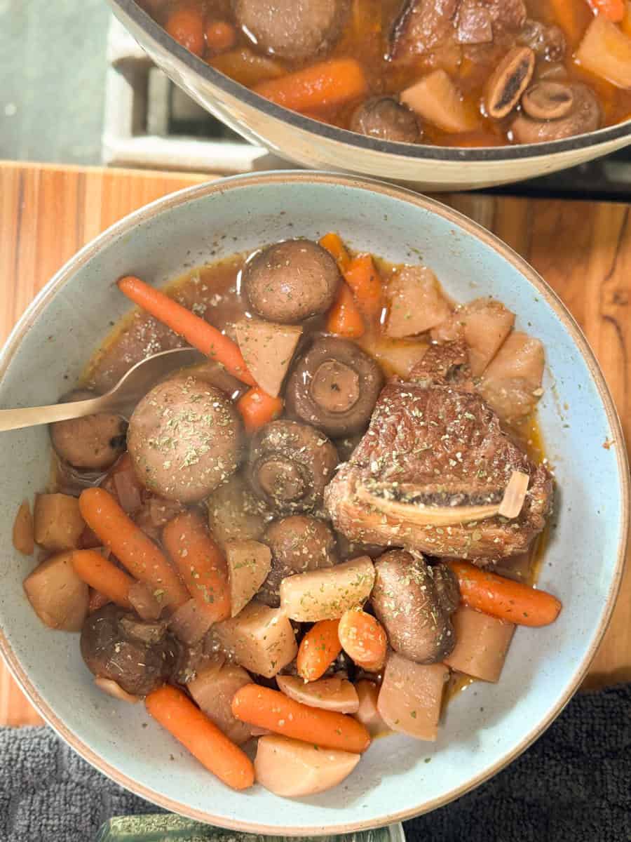 A bowl filled with beef stew, featuring a piece of beef, whole mushrooms, baby carrots, and chunks of potatoes. A spoon rests inside the bowl. In the background, part of a pot with more stew is visible on a wooden surface.