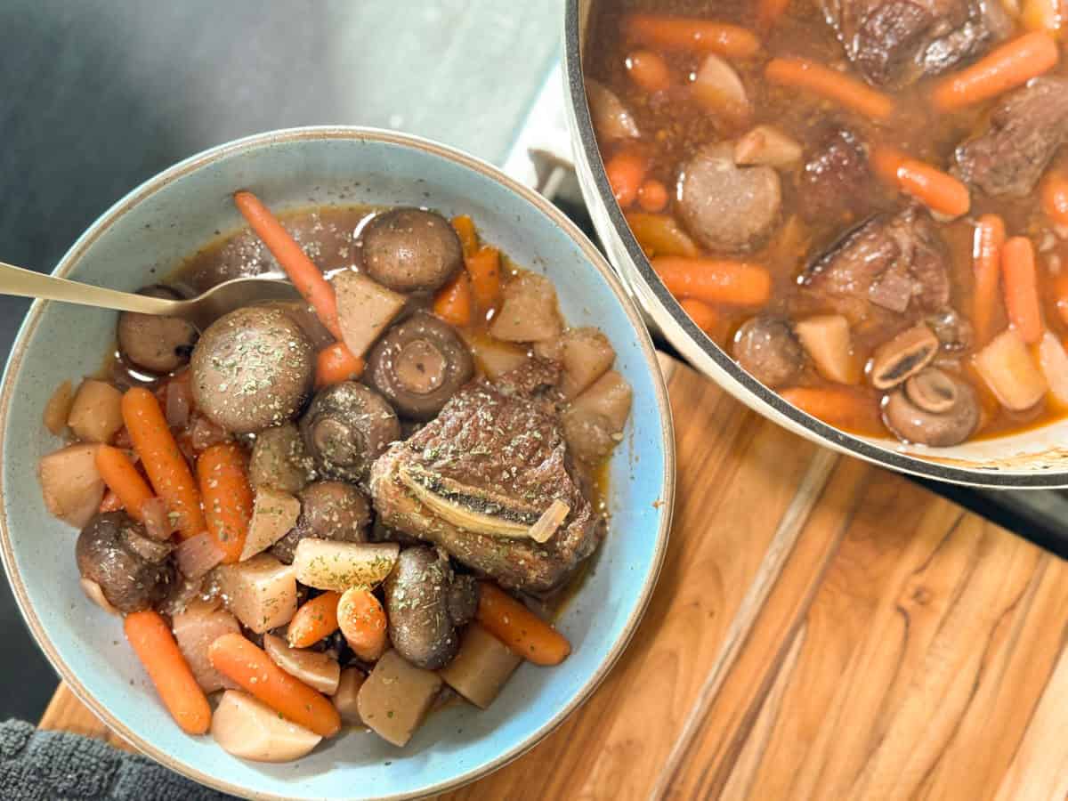 A bowl and a pot of beef stew, filled with chunks of beef, whole mushrooms, baby carrots, and potatoes in a brown broth, are placed on a wooden surface. A spoon is resting in the bowl.
