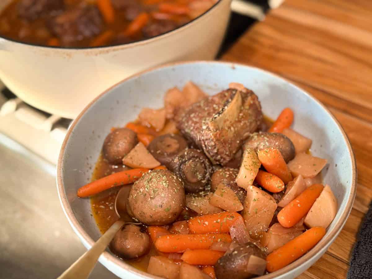 A bowl of beef stew containing beef chunks, whole mushrooms, baby carrots, and diced potatoes. The stew is garnished with herbs and placed on a wooden surface, with a pot of more stew visible in the background.