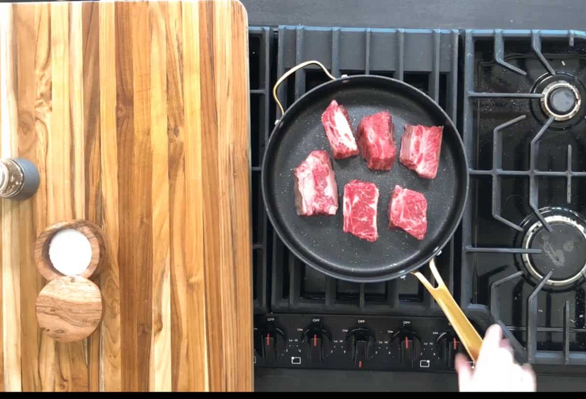 A black frying pan on a stove contains six pieces of raw meat. To the left, there is a wooden cutting board with a small bowl of salt. A hand is holding the pan handle on the right side of the frame.