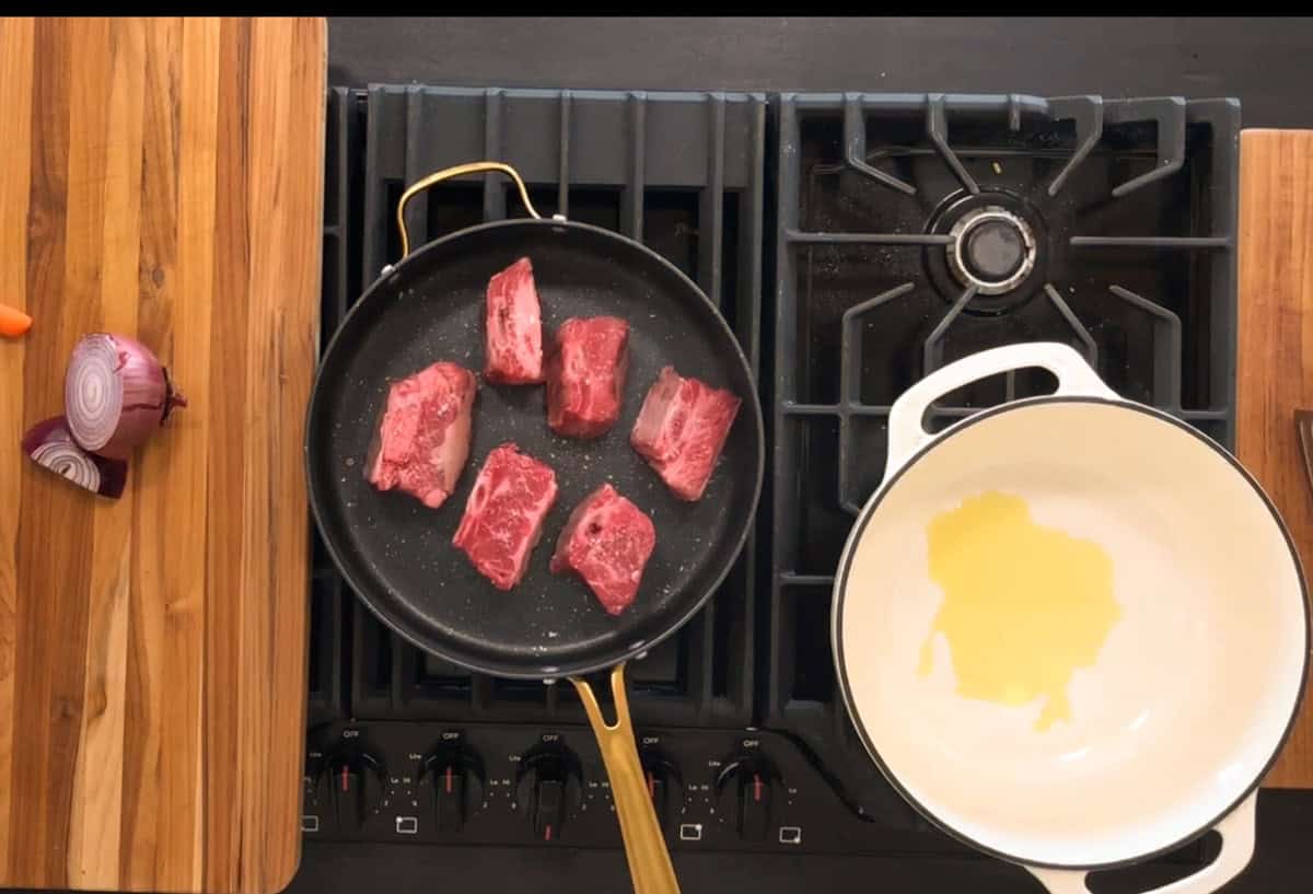A black frying pan with five pieces of raw beef is on a stovetop. Next to it, a white pot with oil is also on the stove. Wooden boards with a sliced red onion and carrot are beside the stove. The scene is set on a kitchen countertop.