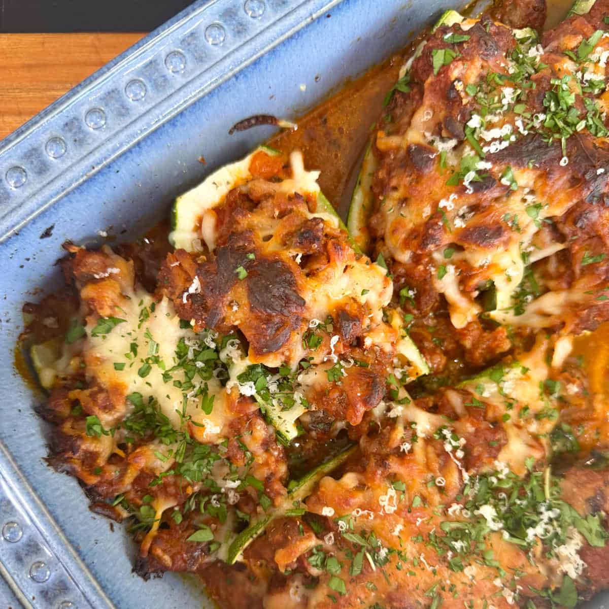 A close-up of stuffed zucchini boats topped with melted cheese and herbs in a blue ceramic baking dish. The boats are filled with a mix of ground meat and tomato sauce, creating a savory, baked dish.