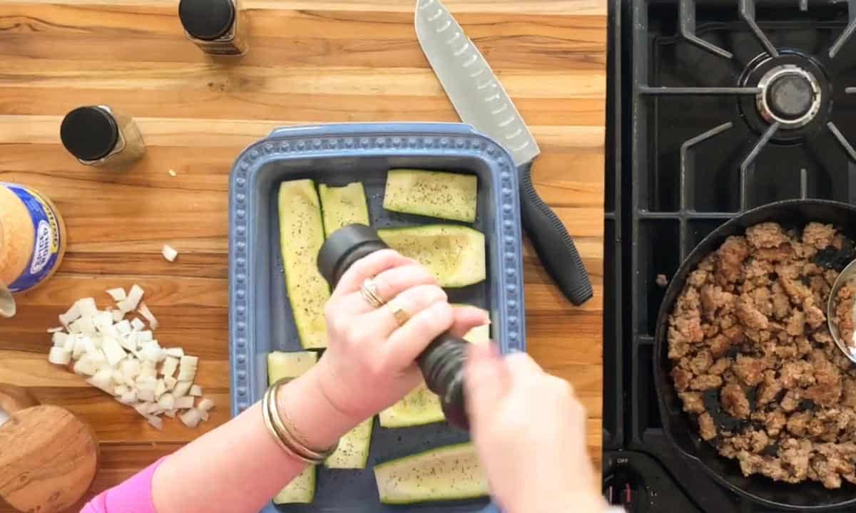 A person seasons sliced green vegetables in a blue baking dish on a wooden countertop. Nearby are chopped onions, spices, a knife, and a skillet with cooked ground meat on a stovetop.