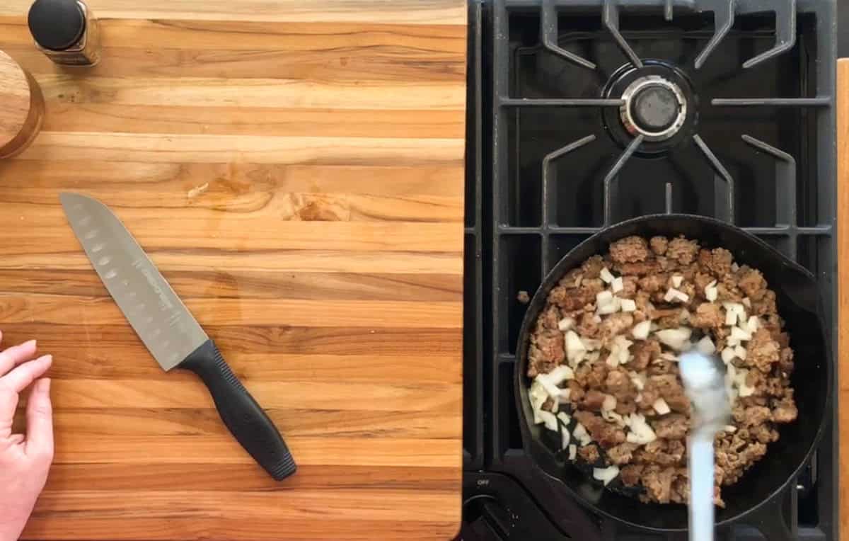 A top-down view of a kitchen scene. On the left, a knife is placed on a wooden countertop. On the right, a person stirs ground meat and diced onions in a black skillet on a gas stove. A seasoning jar is partially visible at the top left.
