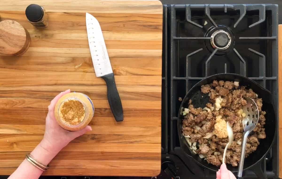 Hand holding a jar of minced garlic over a stovetop. A pan with cooked ground beef and chopped onions is on a gas burner. A slotted spoon rests in the pan. A chef's knife is on the wooden countertop beside two containers.
