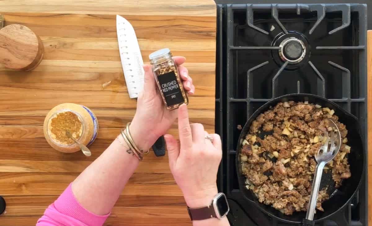 A person is holding a jar of crushed red pepper over a stovetop. There is cooked minced meat with diced onions in a pan on the gas stove. To the left, there's a knife, a jar of garlic, and a small wooden container on a wooden countertop.