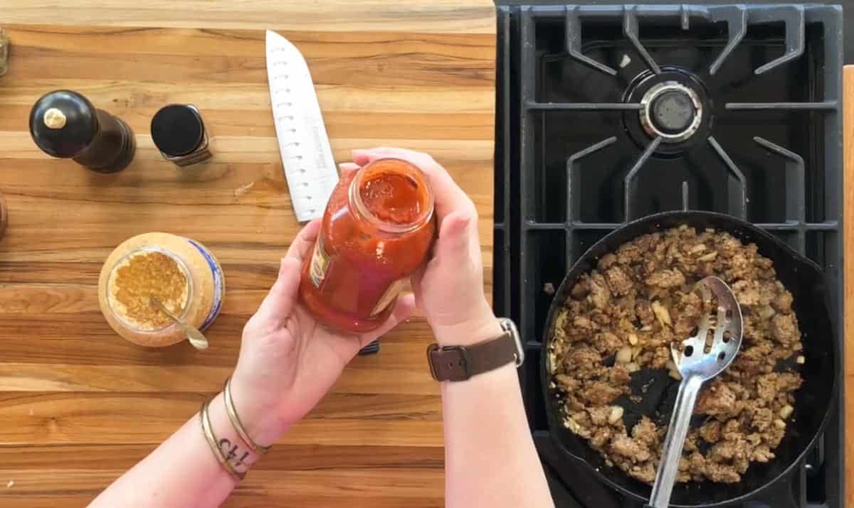 A person holds a jar of tomato sauce above a stove. A pan with browned ground meat is on the burner. On the wooden countertop are a knife, a jar of minced garlic, a pepper mill, and a small container.