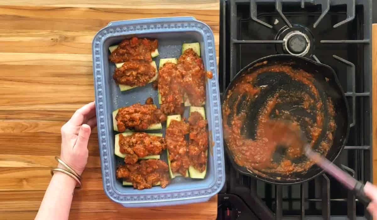 Hands are placing a blue baking dish with stuffed zucchini onto a wooden countertop. The zucchinis are covered with a tomato-based sauce. A black stove is on the right, and a frying pan with sauce residue is to the side.