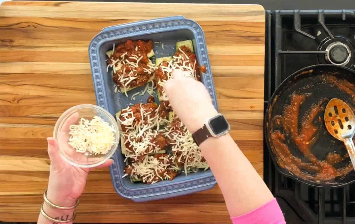 A person sprinkles cheese over a layer of sauce-coated food in a blue rectangular baking dish. The dish is placed on a wooden surface. A pan with sauce remnants and a slotted spoon is on the right. Another bowl of cheese is held in the left hand.