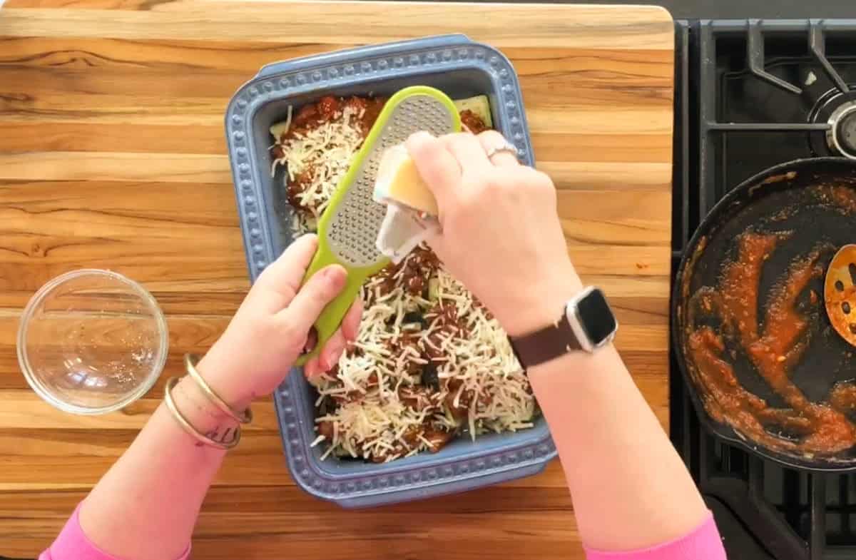Hands are grating cheese over a casserole dish filled with a tomato-based dish in a blue baking pan. A glass bowl is nearby on the wooden countertop, and a pan with sauce residue is on the stove.