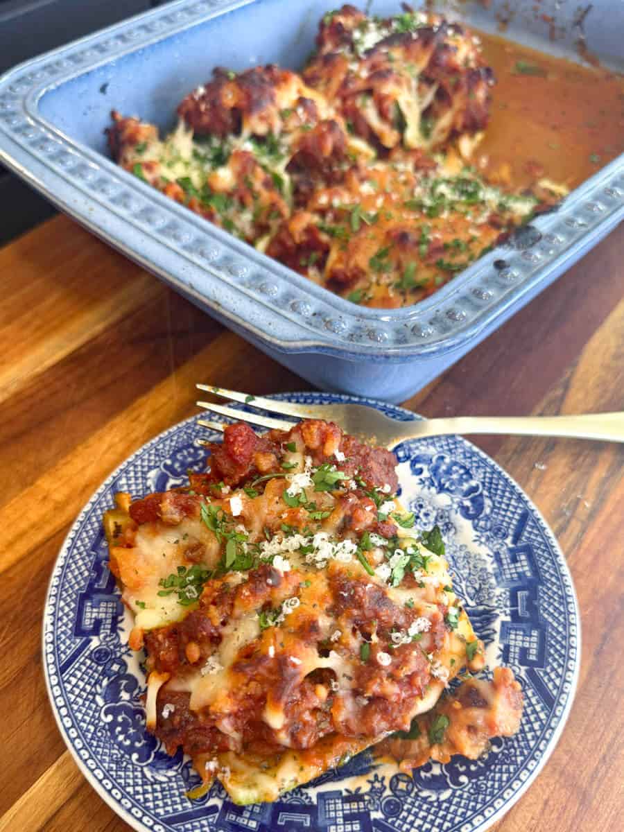 A slice of lasagna on a decorative blue plate, garnished with herbs and grated cheese, sits next to a blue baking dish filled with more lasagna. A fork rests on the wooden surface beside the plate.