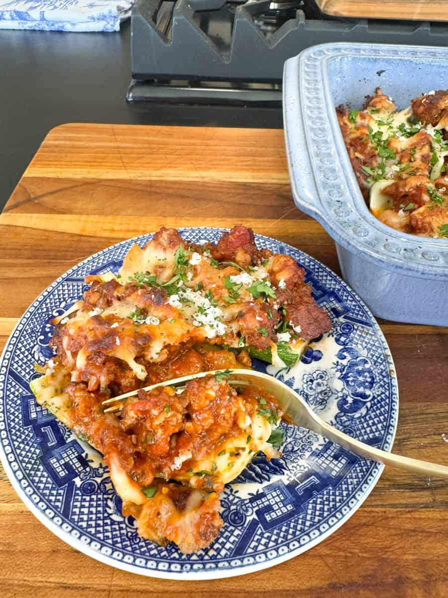 A serving of baked pasta with cheese and meat sauce on a blue and white patterned plate with a fork. A blue ceramic dish containing the rest of the pasta is in the background on a wooden surface. Parsley is sprinkled on top.