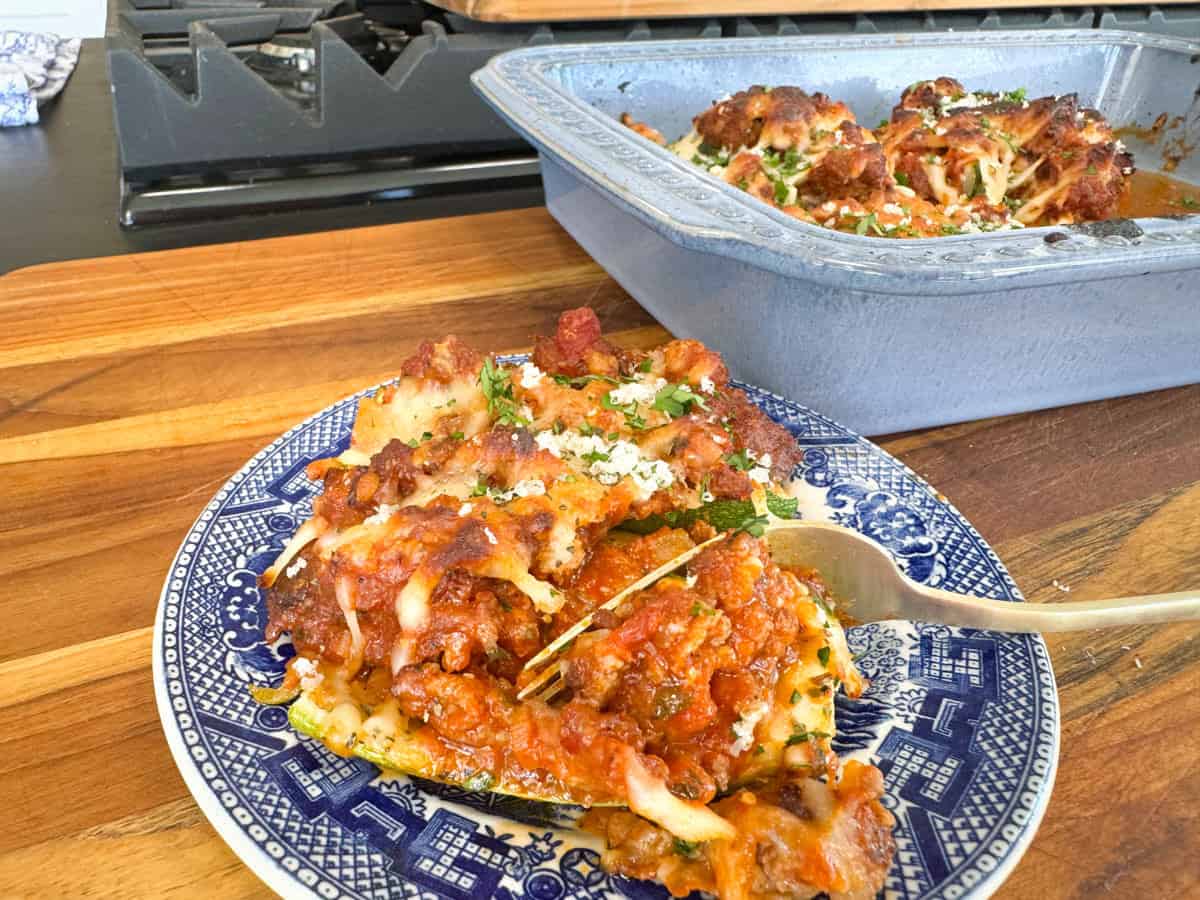 A blue plate with a portion of baked lasagna topped with cheese, placed on a wooden surface. A serving utensil rests on the plate. In the background, a blue baking dish contains the remaining lasagna. A stove is partially visible.