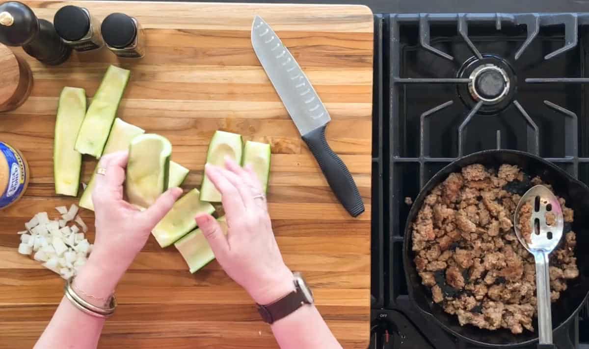 A person prepares a meal with chopped zucchini halves and diced onions on a wooden cutting board. A chef's knife and spice containers are nearby. A skillet on the stove contains cooked ground meat being stirred with a slotted spoon.
