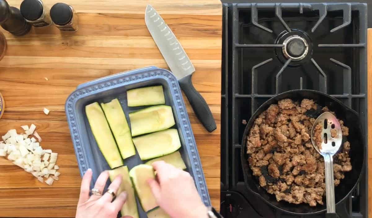 A person prepares zucchini halves in a blue baking dish on a wooden countertop. Chopped onions sit on the side. A frying pan with cooked ground meat is on the stove. A chef's knife lies between the dish and the stove.