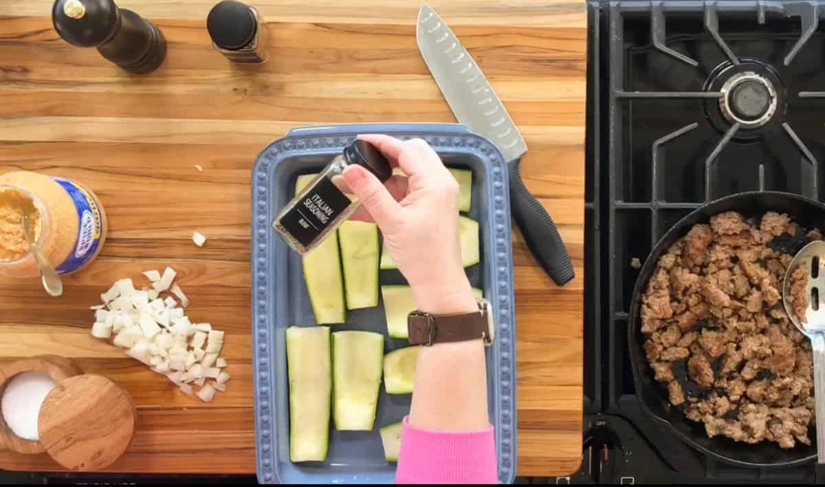 A person holds a bottle of Italian seasoning over a baking dish with halved zucchini. Chopped onions and a jar of peanut butter are nearby. Cooked ground meat is in a pan on the stove, with a knife and a cutting board on the counter.