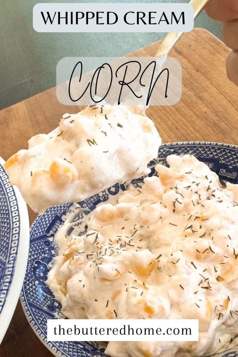A blue and white patterned bowl filled with whipped cream corn garnished with herbs. A spoon holds a portion of the dish above the bowl. The text reads "Whipped Cream Corn" and "thebutteredhome.com.