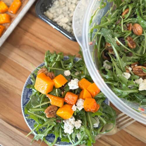 A plate with arugula salad topped with roasted butternut squash, blue cheese, and pecans. A larger container is pouring more salad onto the plate. A pan of squash and a box of blue cheese are in the background on a wooden surface.