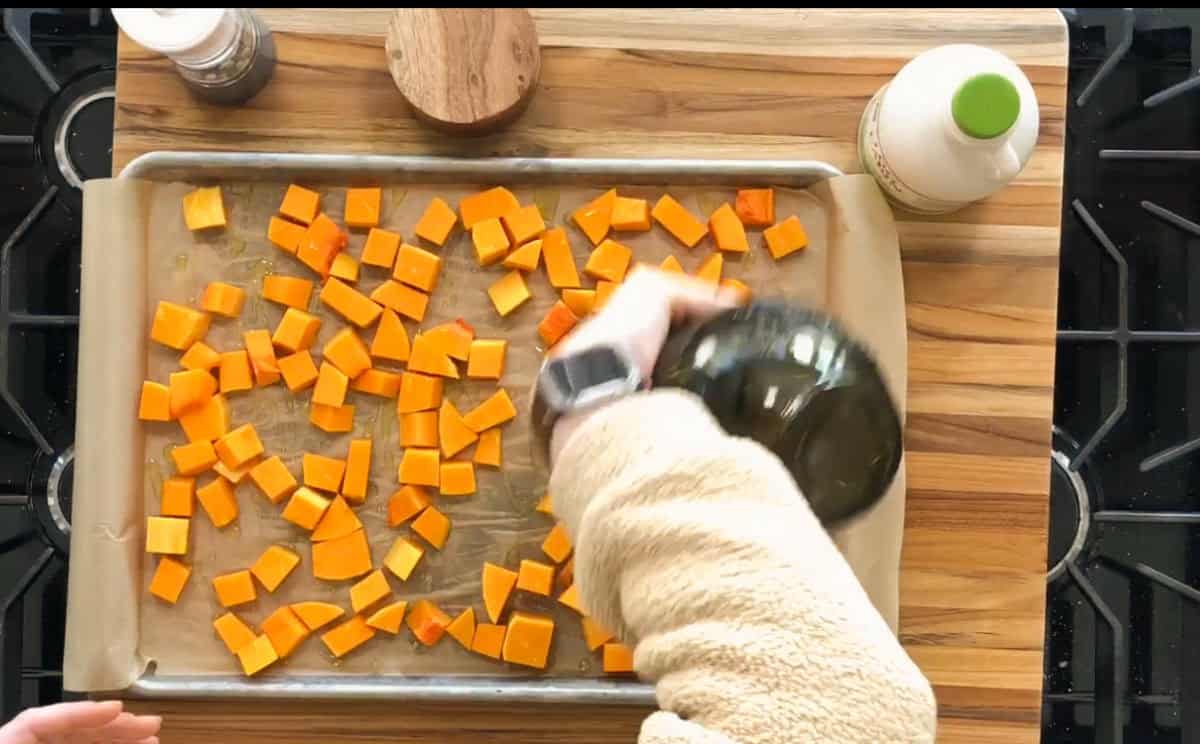 A person wearing a beige sweater drizzles olive oil over cubed orange vegetables, likely butternut squash, spread on a parchment-lined baking sheet. A bottle of olive oil, a pepper grinder, and a bottle of cream are visible nearby on a wooden surface.