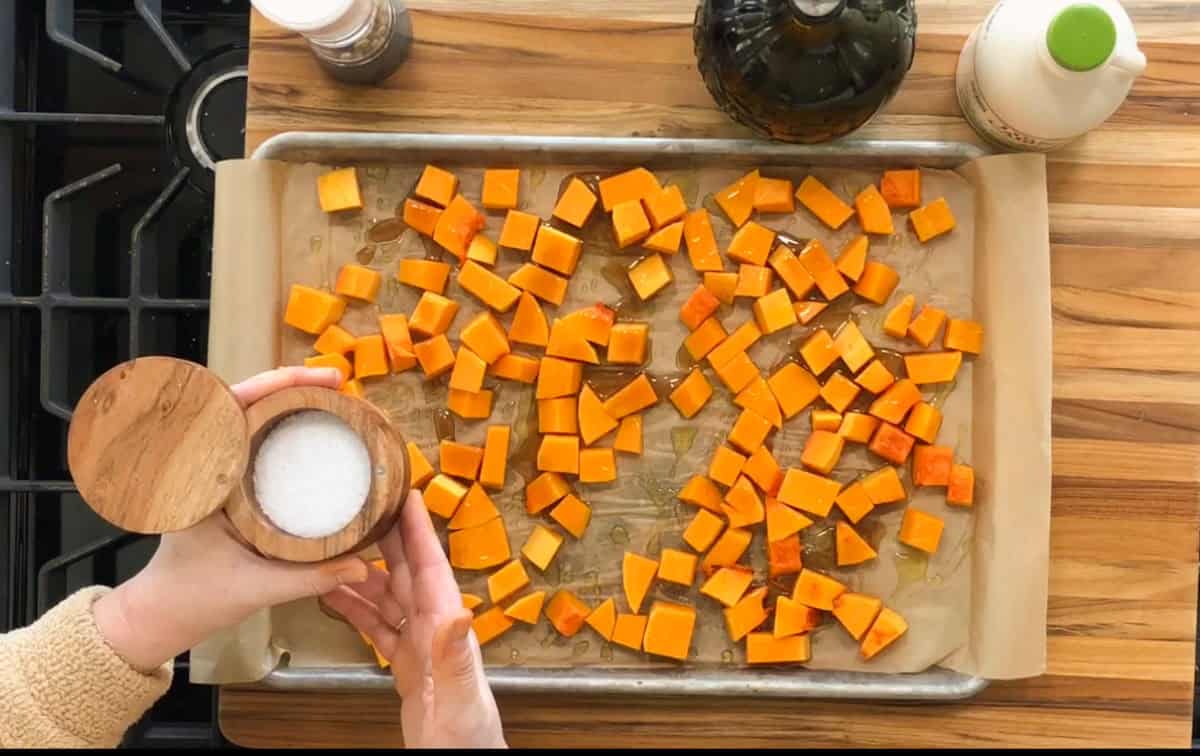 A person is sprinkling salt from a wooden container onto cubed butternut squash spread on a parchment-lined baking tray. The tray is on a wooden countertop next to a bottle of oil and a spice grinder.