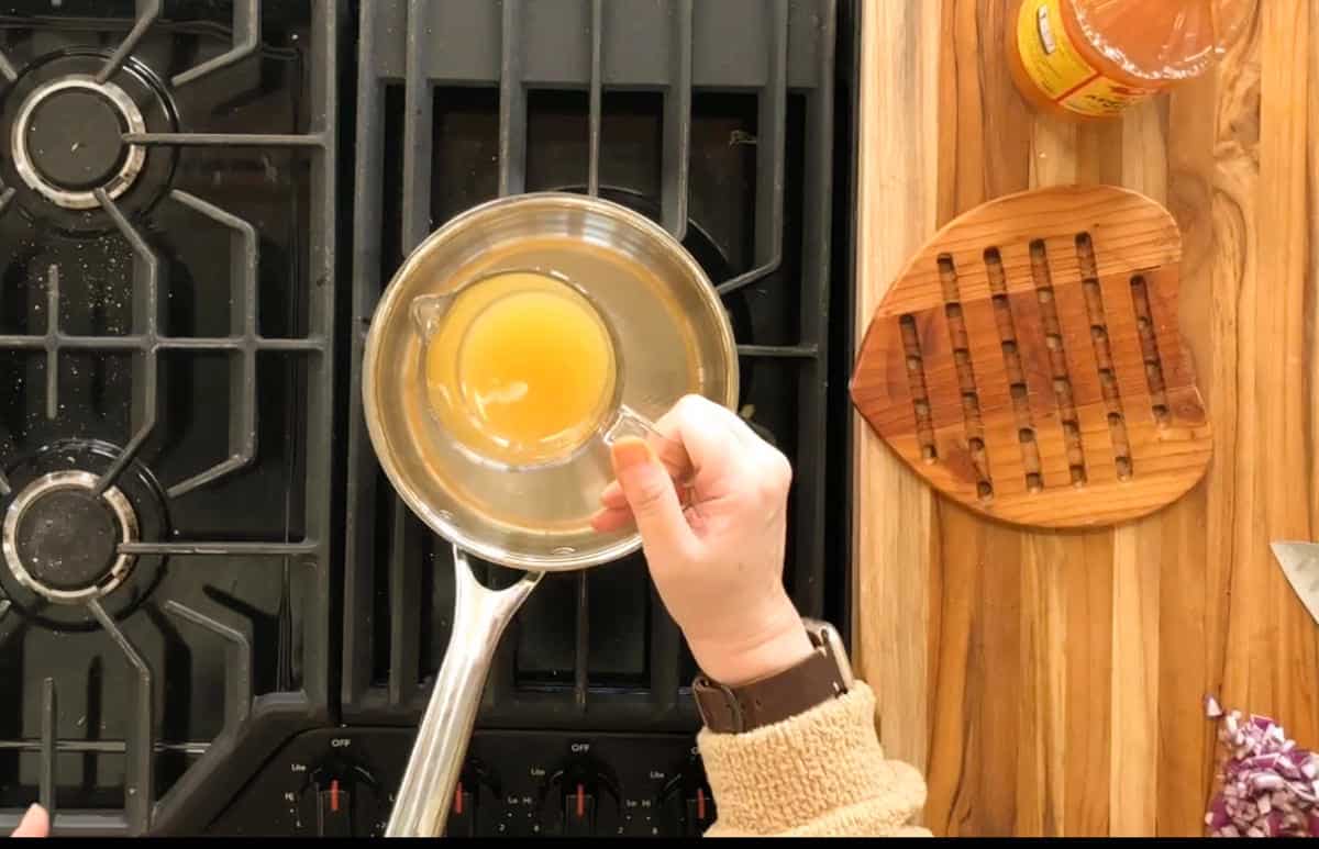 A person wearing a tan sweater holds a measuring cup of orange liquid over a saucepan on a stove. The countertop has a bottle, a wooden trivet shaped like a heart, and chopped red onion.