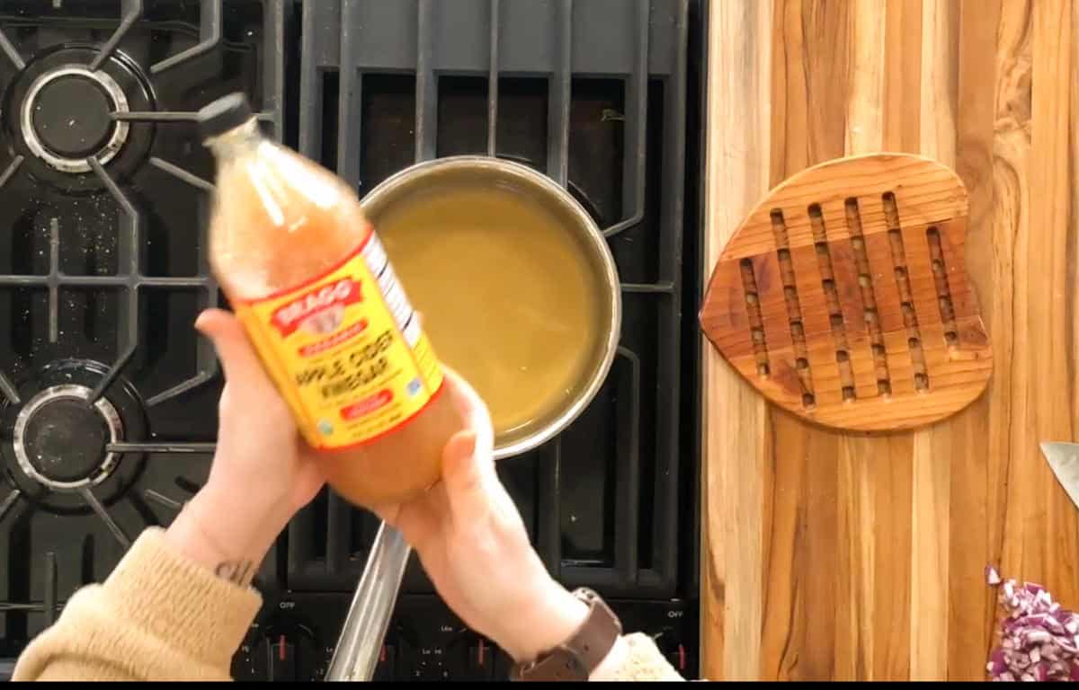 A person holds a bottle of apple cider vinegar over a saucepan on a stove. There is a wooden cutting board with a groove pattern on the right and a small pile of chopped vegetables on the bottom right corner.