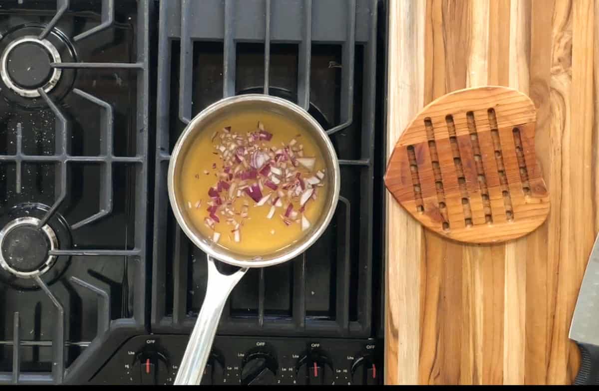 A saucepan is on a stove with a black grate, containing liquid and chopped red onions. To the right, there is a wooden chopping board with a handle, placed on a wooden countertop.