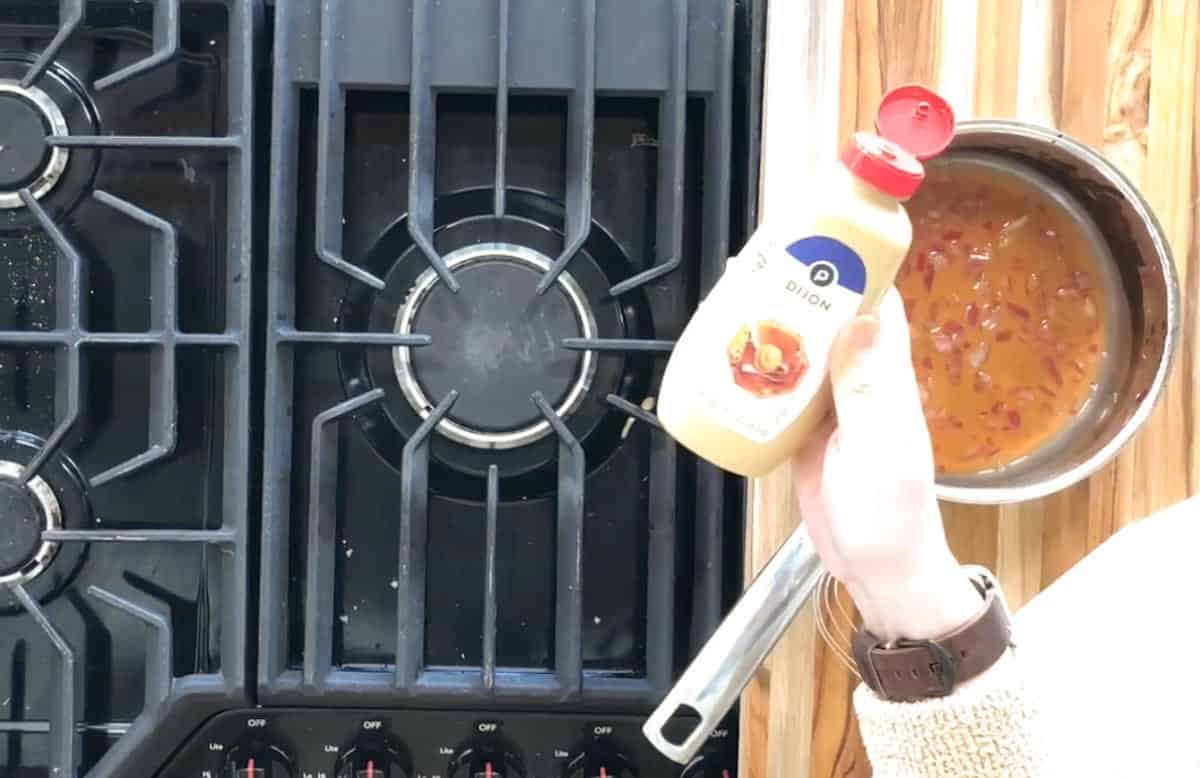 A hand holds a bottle of Dijon mustard over a saucepan containing a red sauce, positioned on a wooden countertop beside a gas stove with four burners.