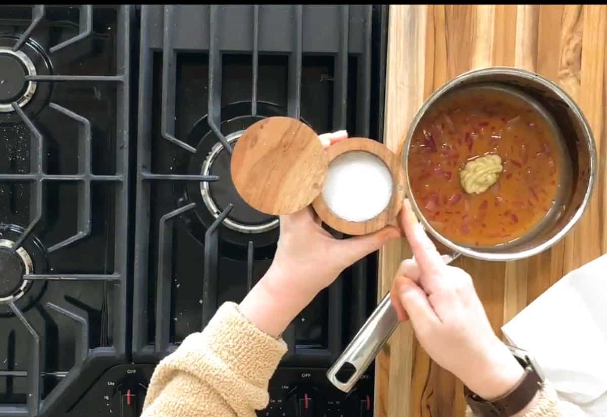 Person holding a wooden salt container above a saucepan with red sauce and a dollop of mustard on a stovetop. The stove has black grates, and a wooden cutting board is visible on the right. The person's hand is about to add salt to the pan.