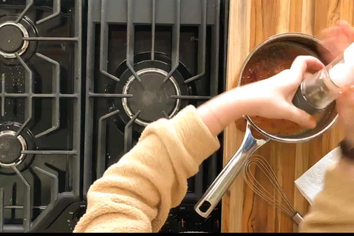 A person wearing a beige sweater is grinding pepper or seasoning into a pot on a wooden countertop. There is a metal whisk nearby, and the pot is next to a stove with visible burners.