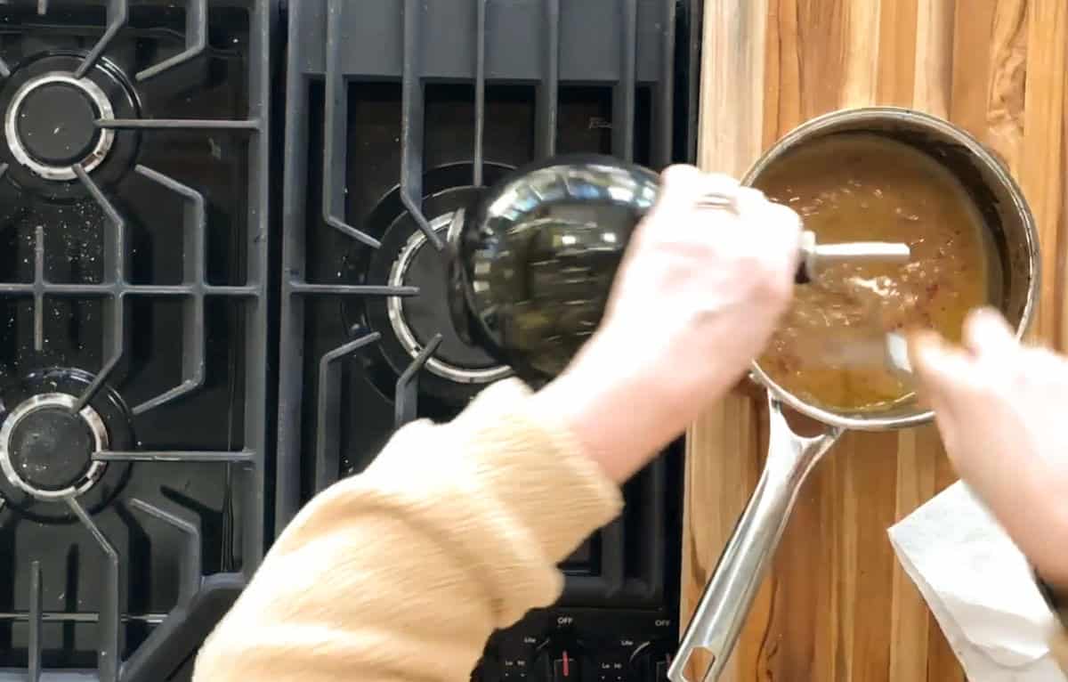 A person uses an immersion blender in a saucepan filled with a brown liquid on a wooden countertop. The stovetop features gas burners on the left. The person wears a beige sweater and a ring. A white paper is partially visible on the counter.