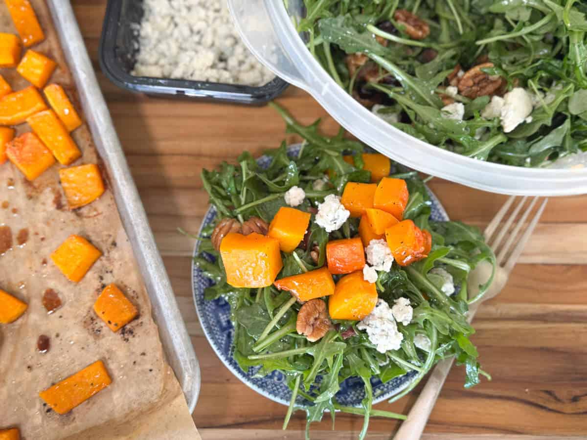 A blue plate with an arugula salad topped with roasted butternut squash, pecans, and crumbled cheese. A baking sheet with more squash and a container with additional salad are nearby. A fork is placed beside the plate.