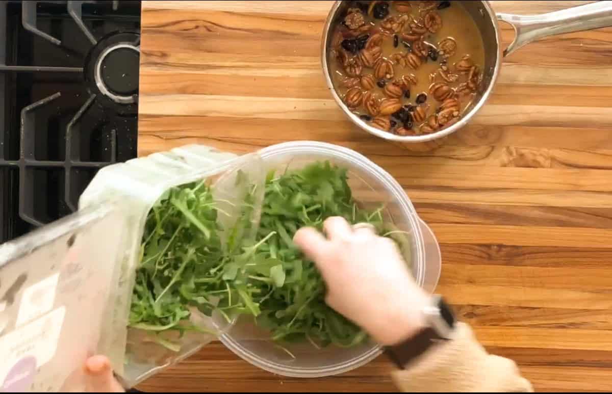 A person is placing arugula into a clear bowl from a plastic container on a wooden countertop. A saucepan containing pecans and dried fruit is nearby. A section of a gas stovetop is visible on the left.