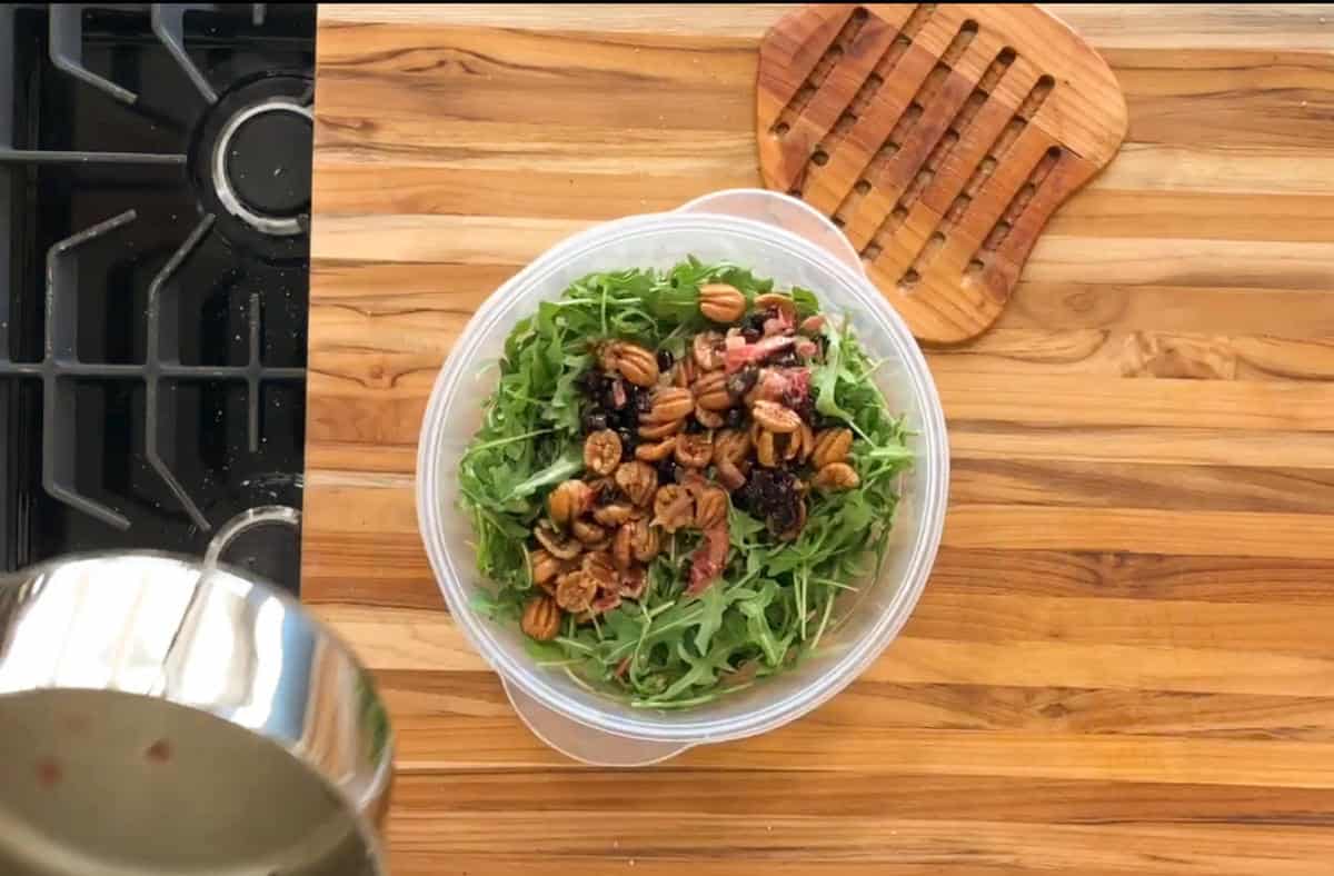 A bowl of salad with arugula, pecans, and sliced meat, placed on a wooden countertop. A pot is visible on the left side near a stove, with a trivet in the upper right corner of the counter.