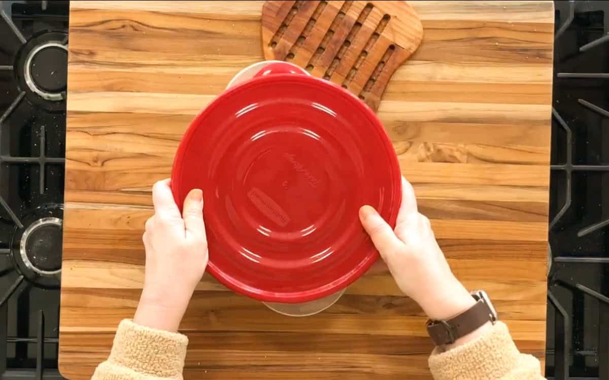 A person holds a red lid over a round glass dish on a wooden cutting board. The stove burners are visible on either side. A wooden trivet is placed nearby on the board. The person wears a brown watch and a beige fleece sweater.