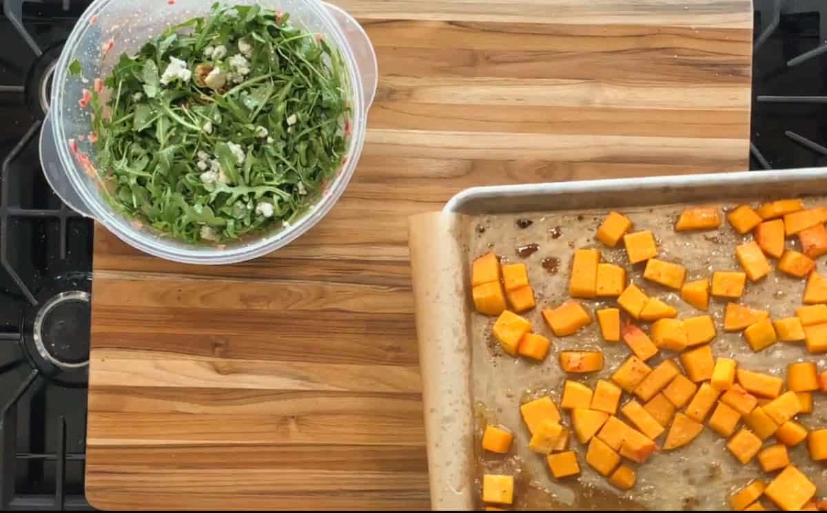 A wooden countertop holds a metal baking sheet with parchment paper and cubed butternut squash. Next to it is a bowl of arugula salad mixed with crumbled cheese and nuts.