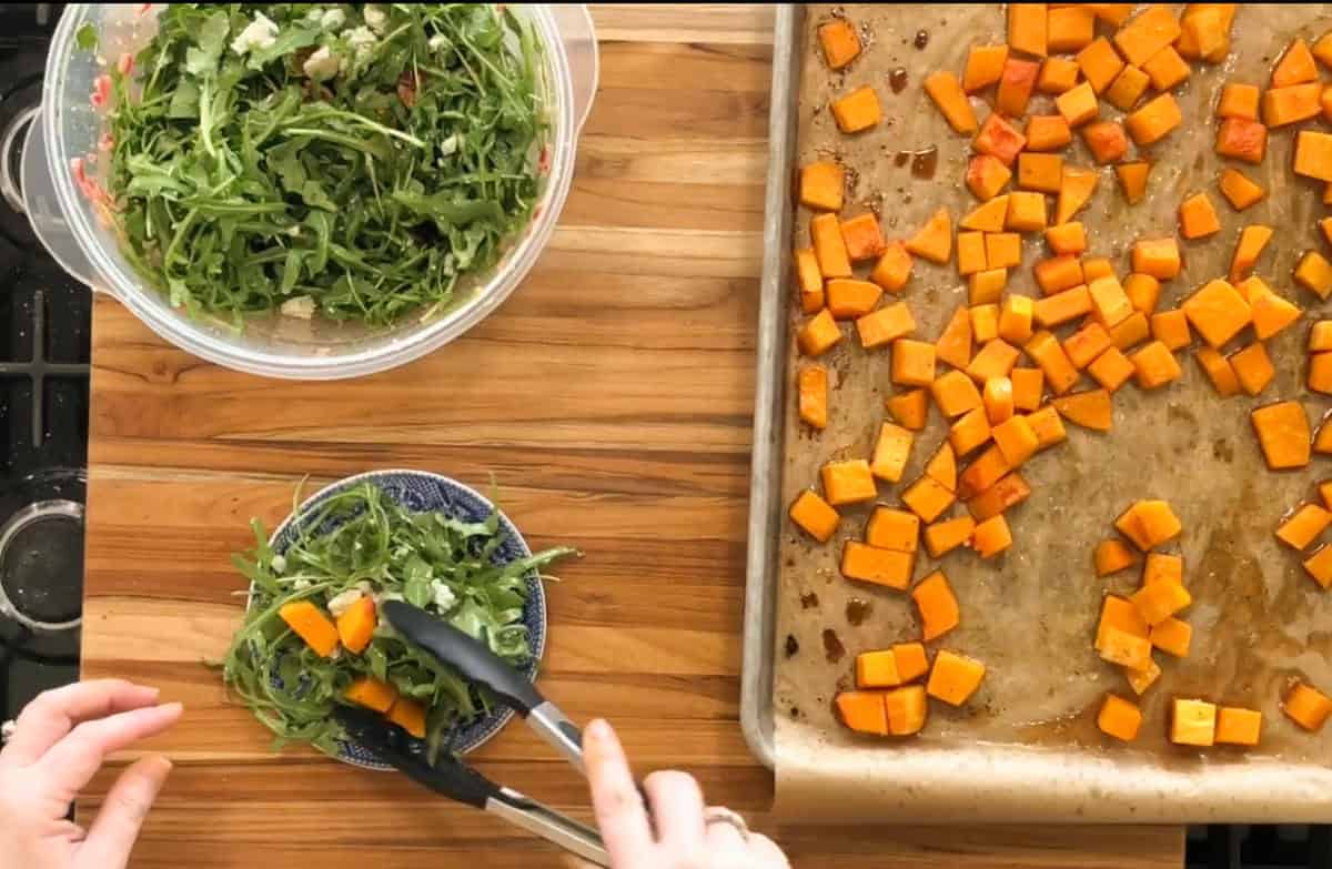 A wooden table with a baking sheet of roasted butternut squash cubes on the right. A bowl of arugula salad is on the left, and a hand uses tongs to add squash to a smaller bowl of salad.