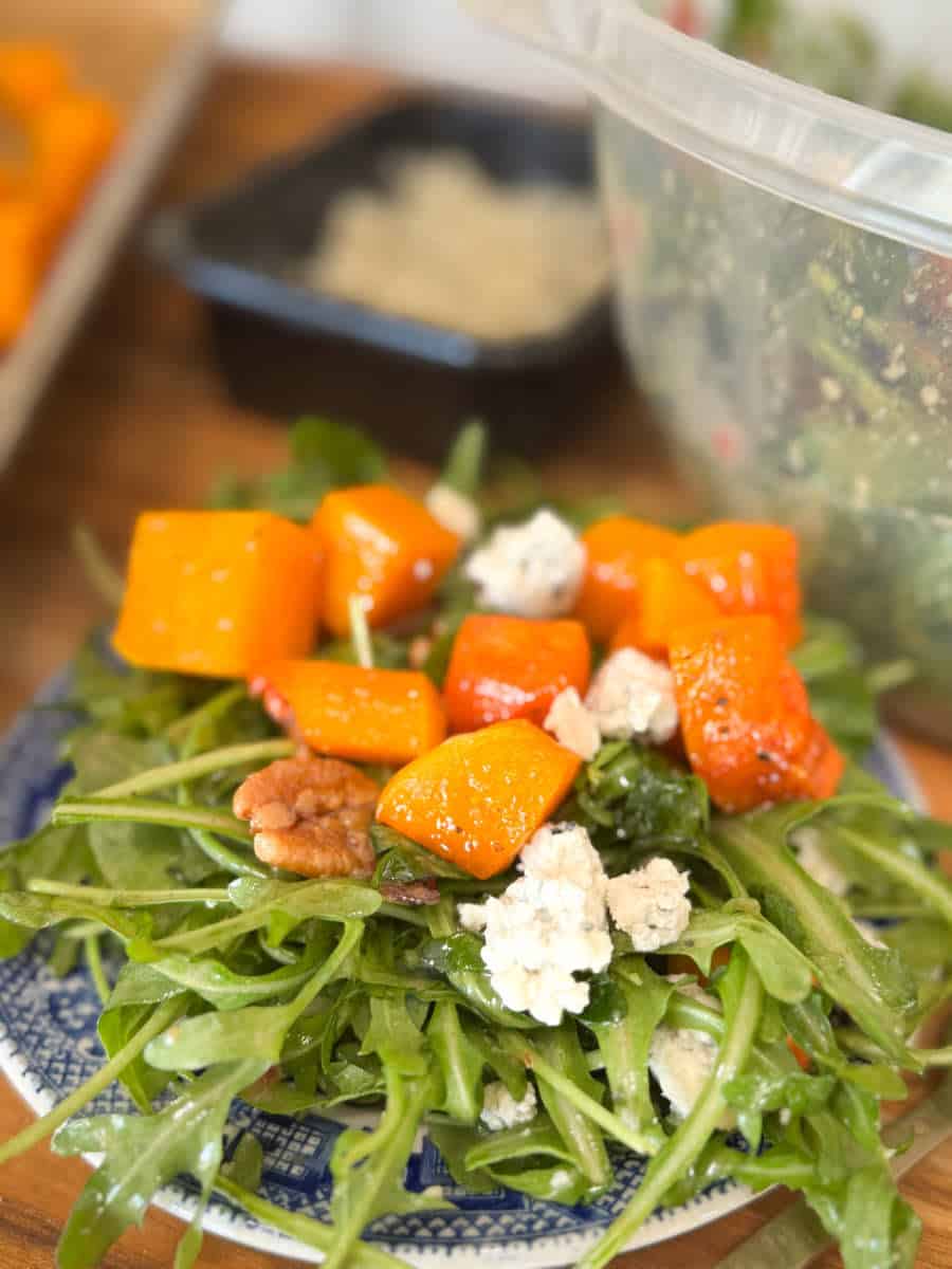 A salad on a blue plate features arugula, roasted butternut squash cubes, walnuts, and crumbled blue cheese, with a plastic container of salad visible in the background.